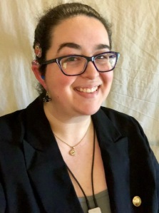 A dark haired woman, with hair pulled back and dark-framed glasses is smiling. She wears a dark colored blazer and has a cochlear implant.