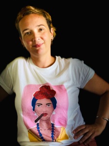 A white woman with light brown short hair smiles with her hand on her hip. She is wearing a white t-shirt with an image of Frida Kahlo.