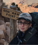 Young white woman sites with a canyon behind her. She wears a baseball hat and glasses. The wooden  sign next to her says "Ooh Aah Point".