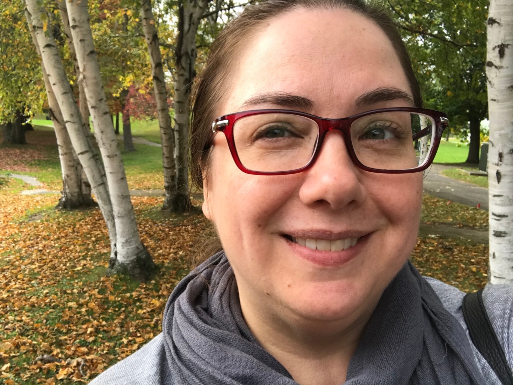 White woman smiles with dark hair pulled back and red rimmed glasses. She is looking to the side of the camera. Behind her are birch trees and autumn leaves on the ground.