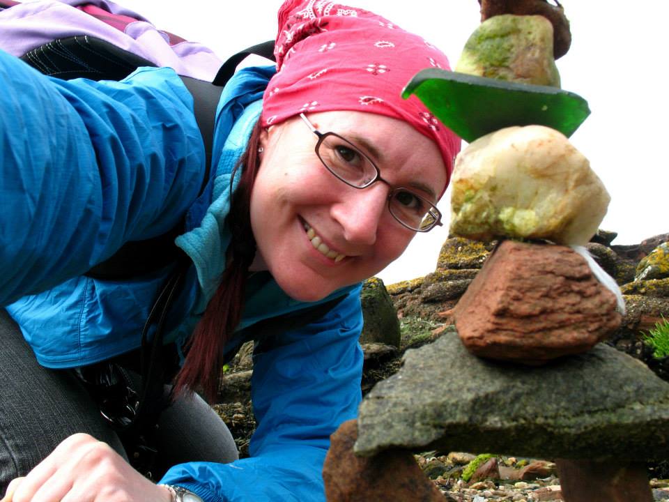 White woman with bandana around her hair leans near the ground just behind a stack of rocks and sea glass. She wears glasses and outdoor gear.