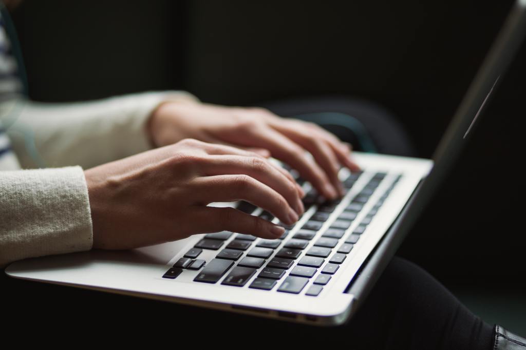 Feminine hands top on a laptop. The typer wears an off-white sweater. Photo by Kaitlyn Baker on Unsplash.
  