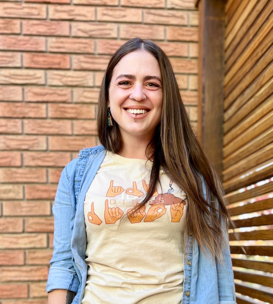 woman with long brown hair and pale skin smiles to the camera wearing a beige shirt that says in fingerspelling font "IDK DINOS".  The shirt is from Amelia's shop.