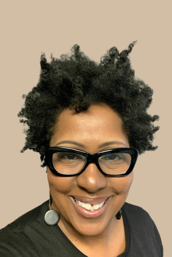Dark skinned woman with stylish glasses smiles to the camera. She wears a black shirt and dangly earrings.
